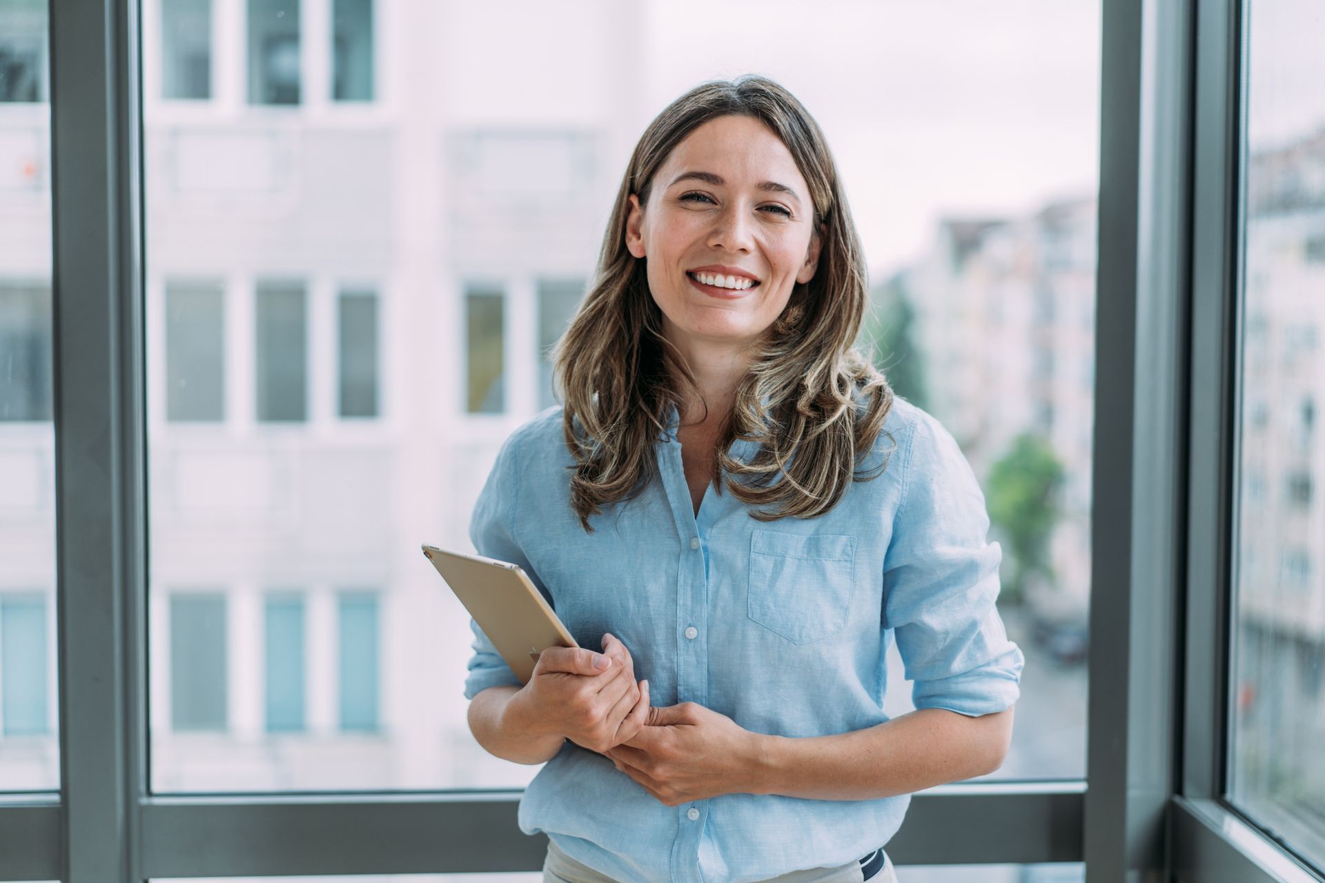 A woman is standing in front of a window holding a tablet and smiling.