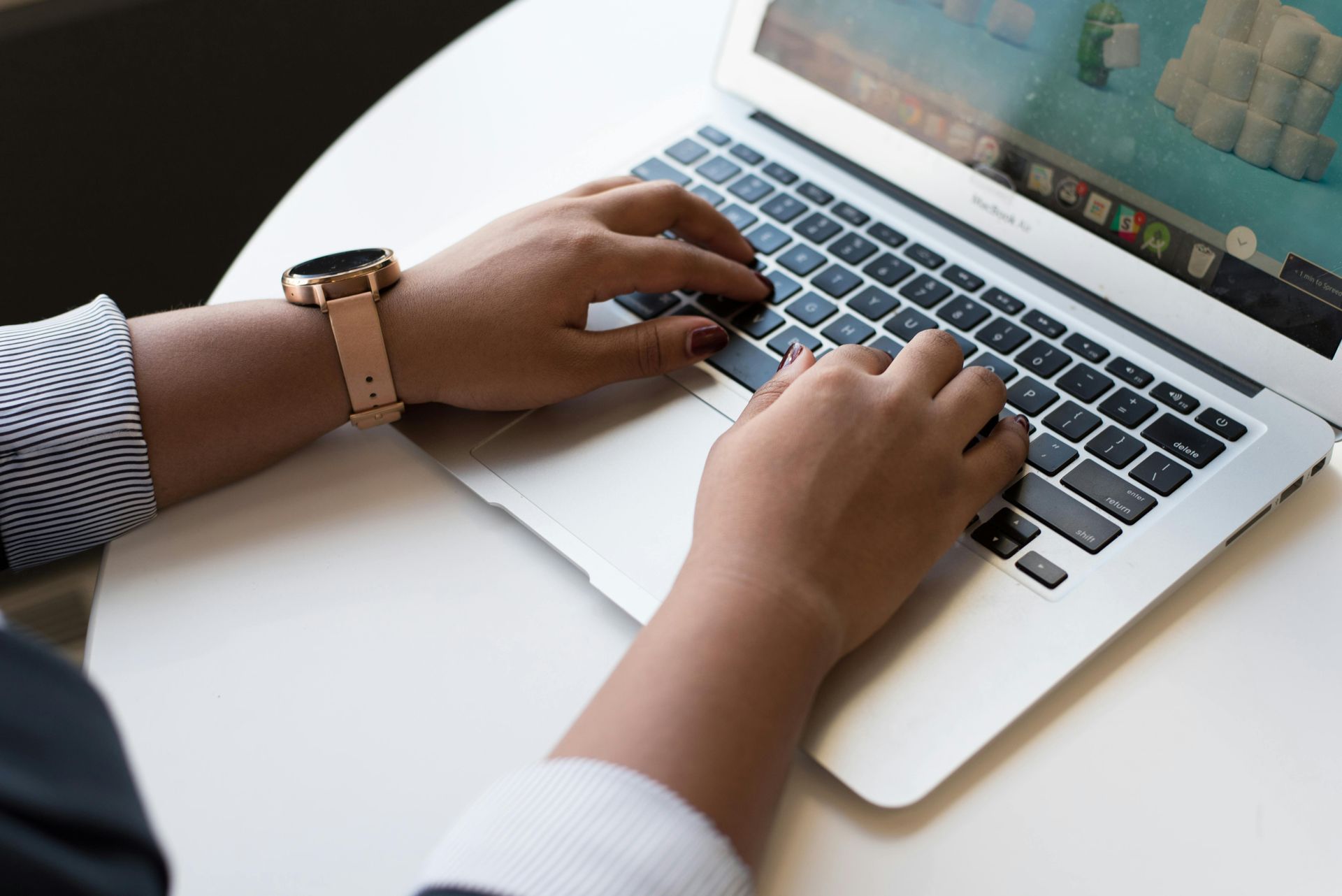 Hands typing on a laptop with a watch on one wrist, angled on a white table.