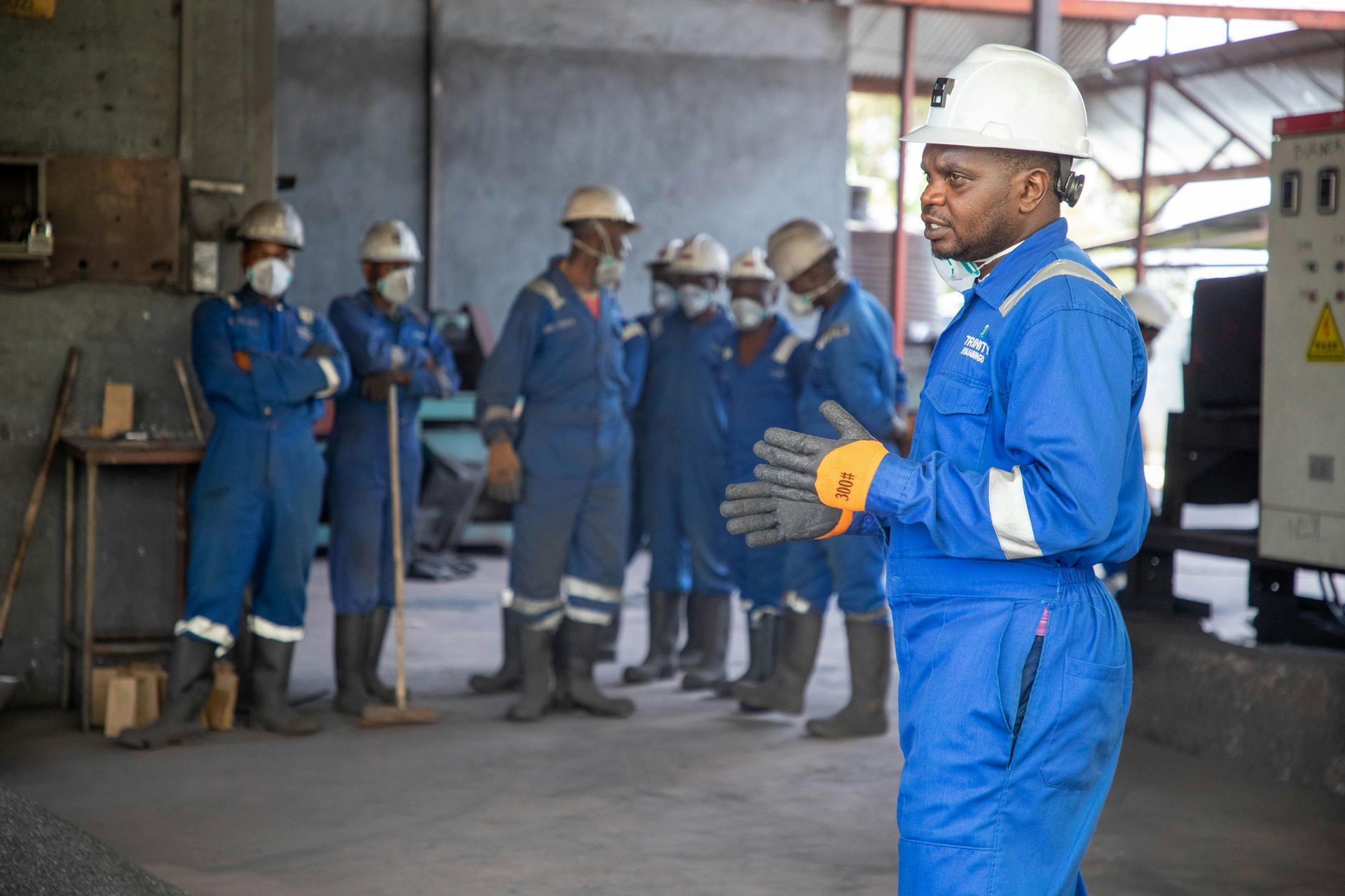 Man in blue coveralls and hard hat speaks to a group of workers in an industrial setting.