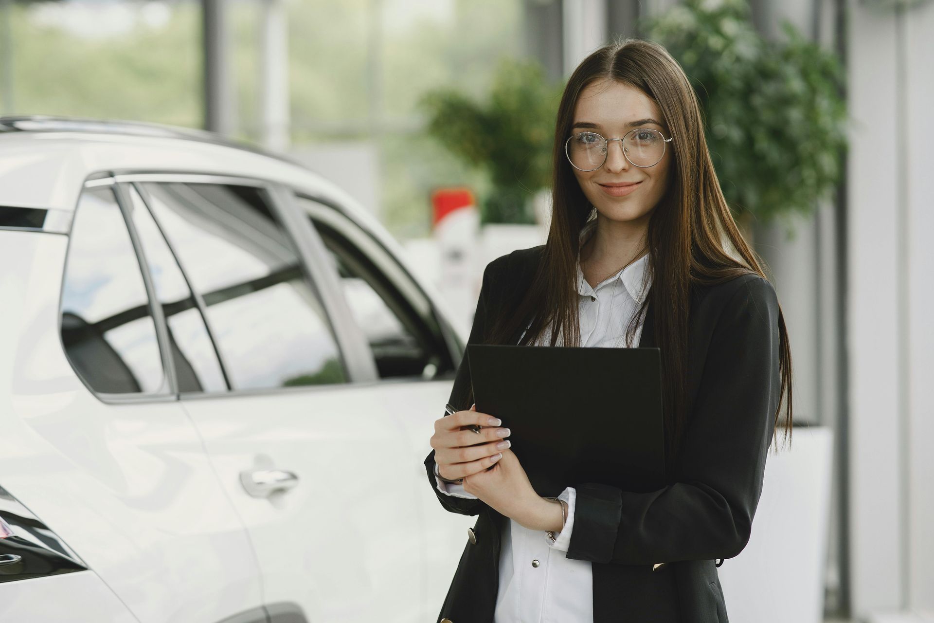 Woman in a blazer holds a tablet, smiling, standing near a white car in a showroom.