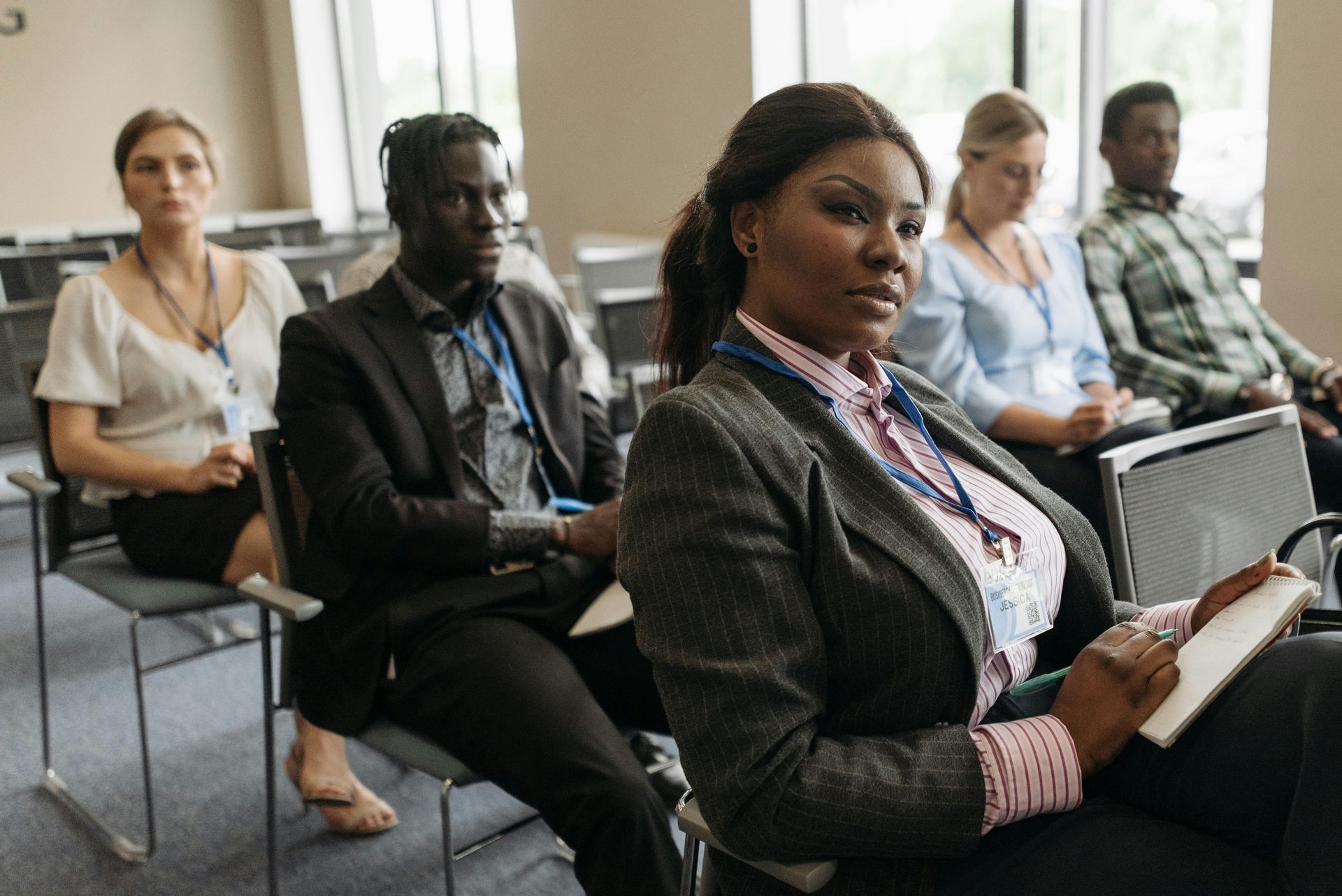 People in business attire at a conference, seated and listening, some holding notebooks.
