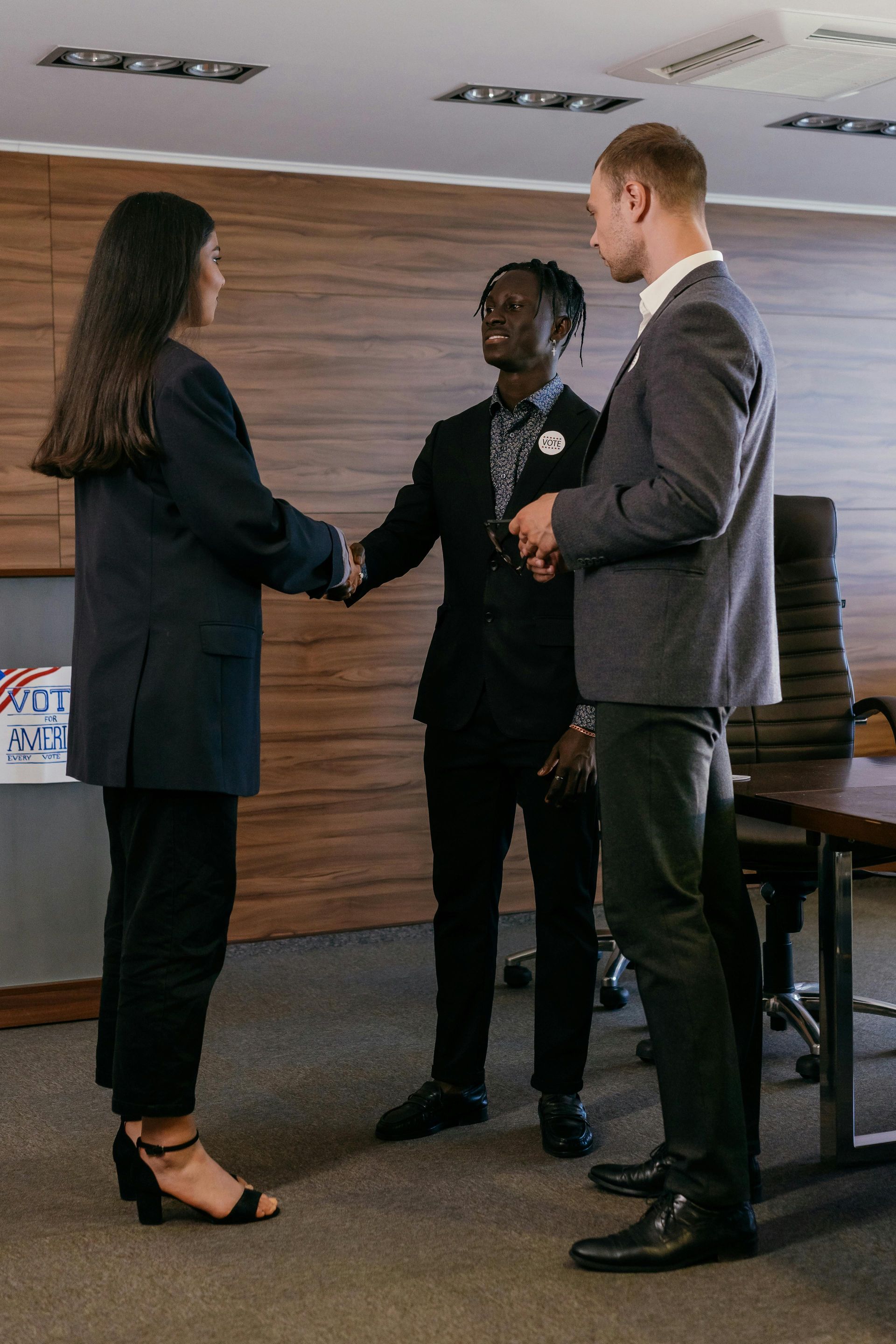 Three people in business suits, one shaking hands. Office setting with wooden paneling.