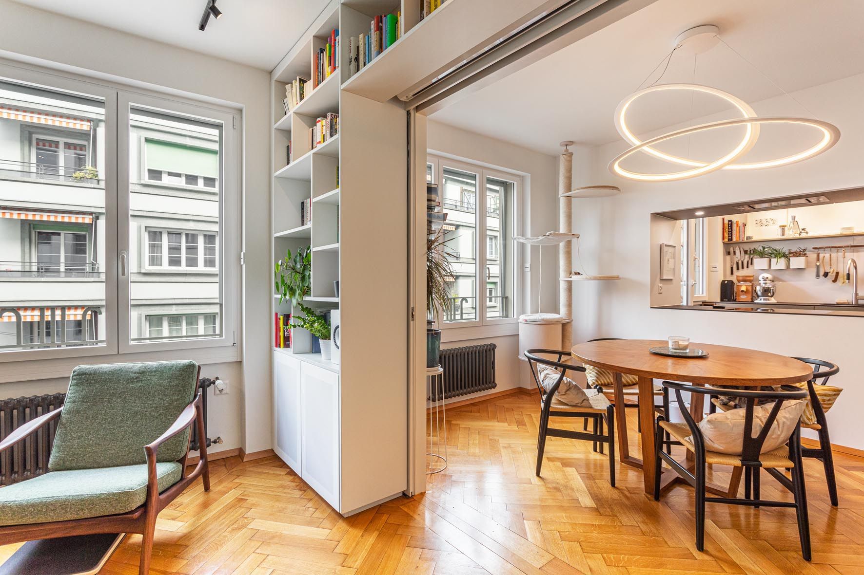 Appartement moderne et lumineux avec table à manger, suspension lumineuse et étagères ouvertes près d'une banquette.