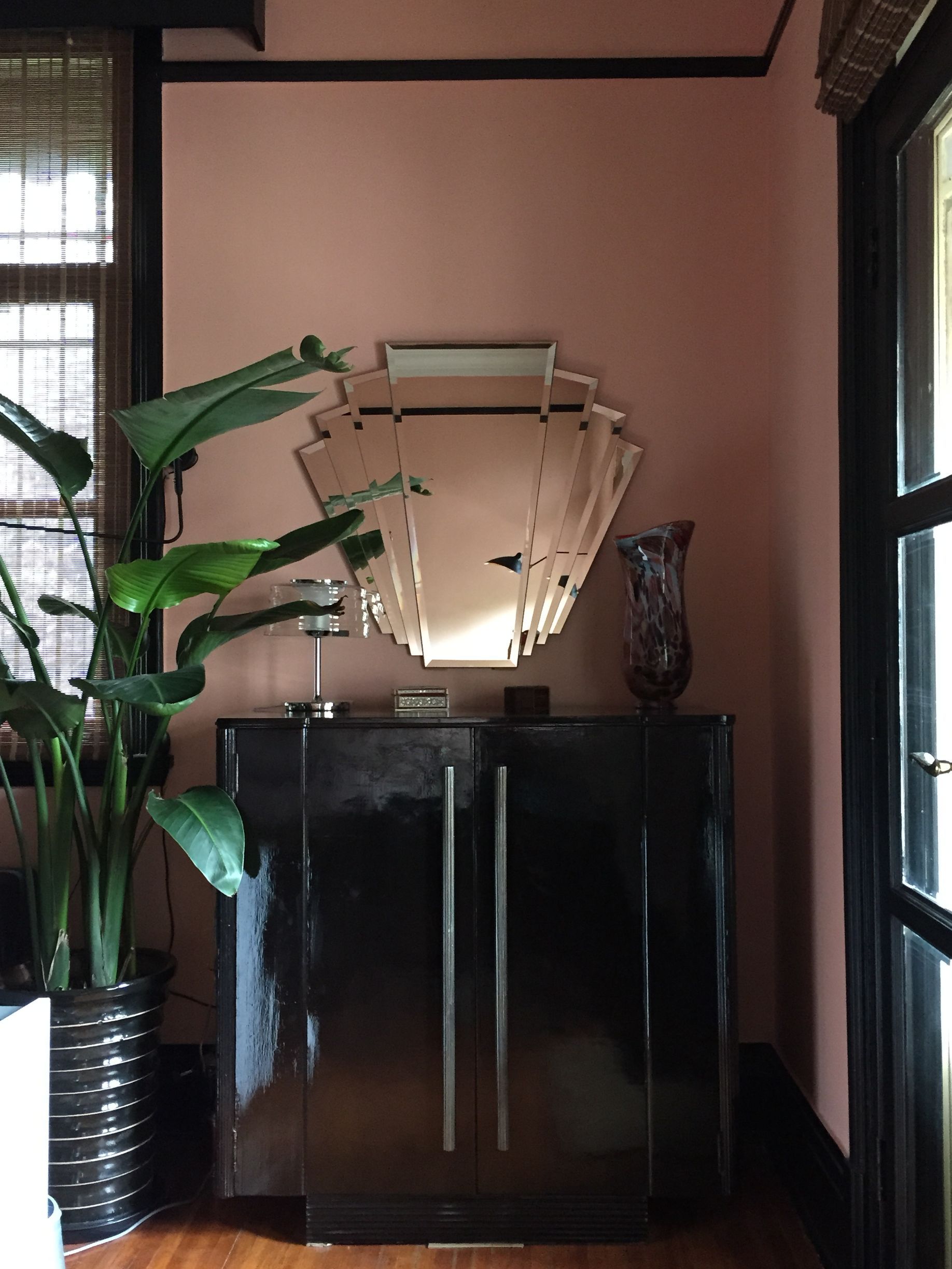 Console moderne avec miroir décoratif dans une pièce aux murs roses, encadrée par des plantes et une fenêtre