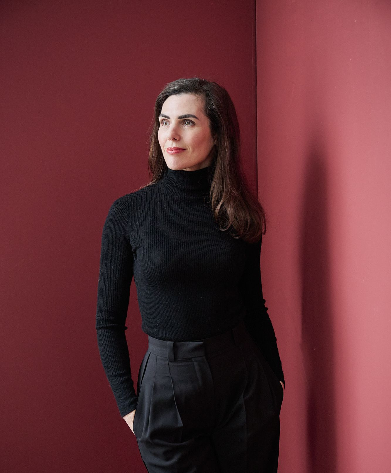 Femme en col roulé noir et pantalon, debout contre un mur rouge, regardant sur le côté.