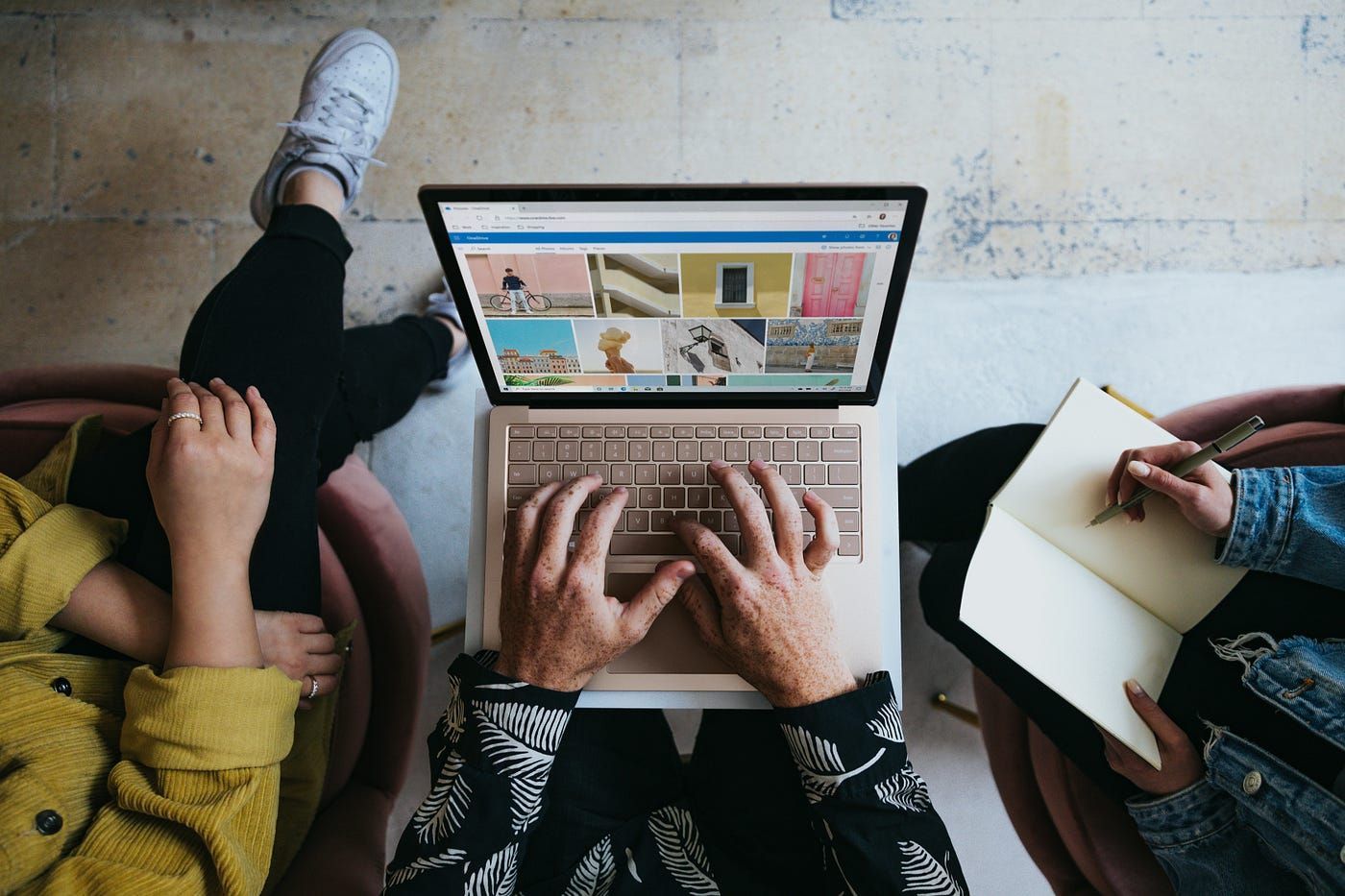 Hands typing on laptop, two others nearby; one writing in notebook, the other's legs visible.