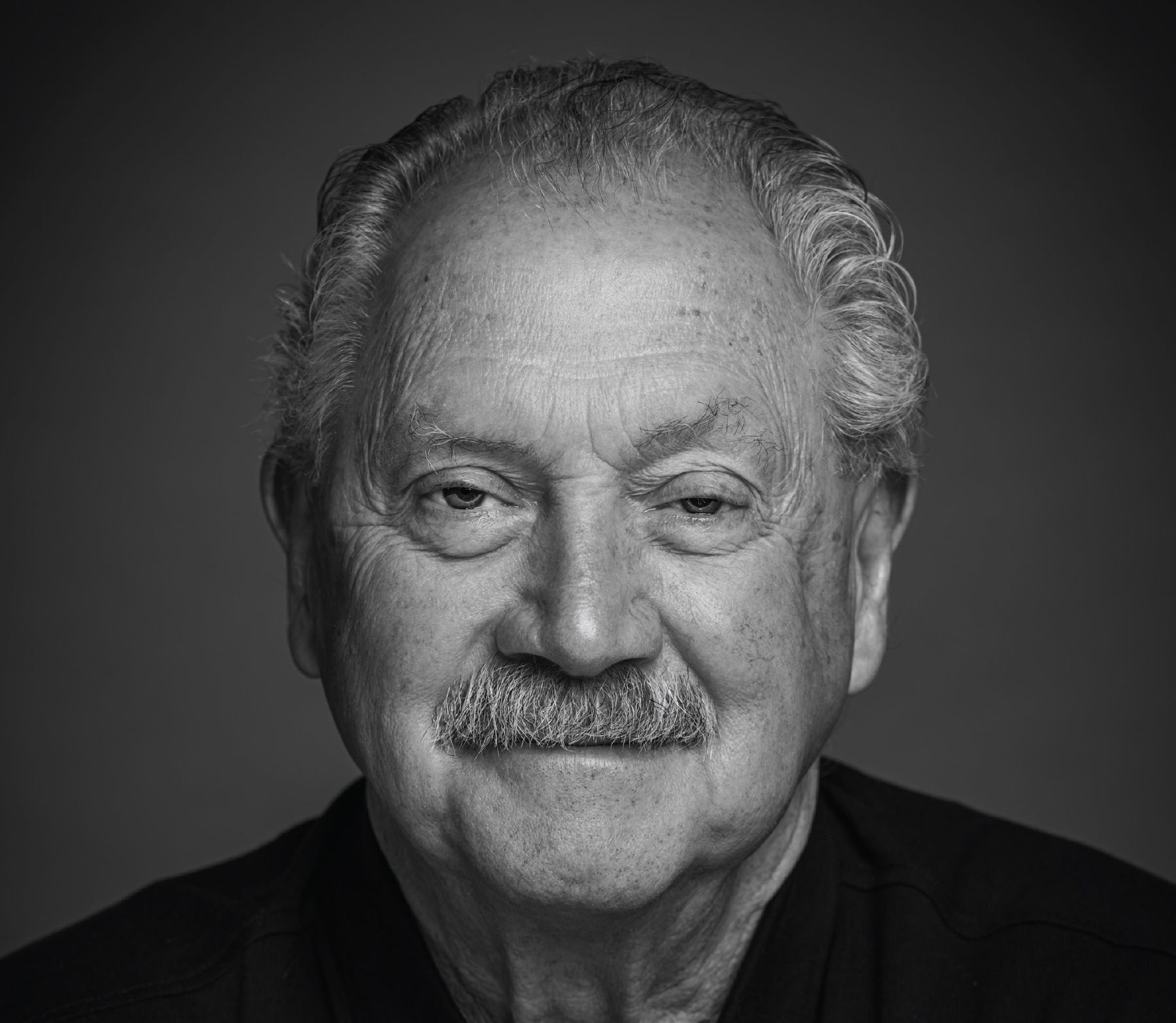 Black and white portrait of an older man with a mustache, looking directly at the camera, smiling slightly.