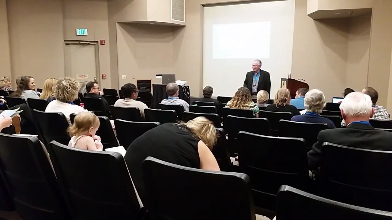 A man speaks at a podium to an audience in a conference room. People sit in rows of chairs, listening to the speaker.