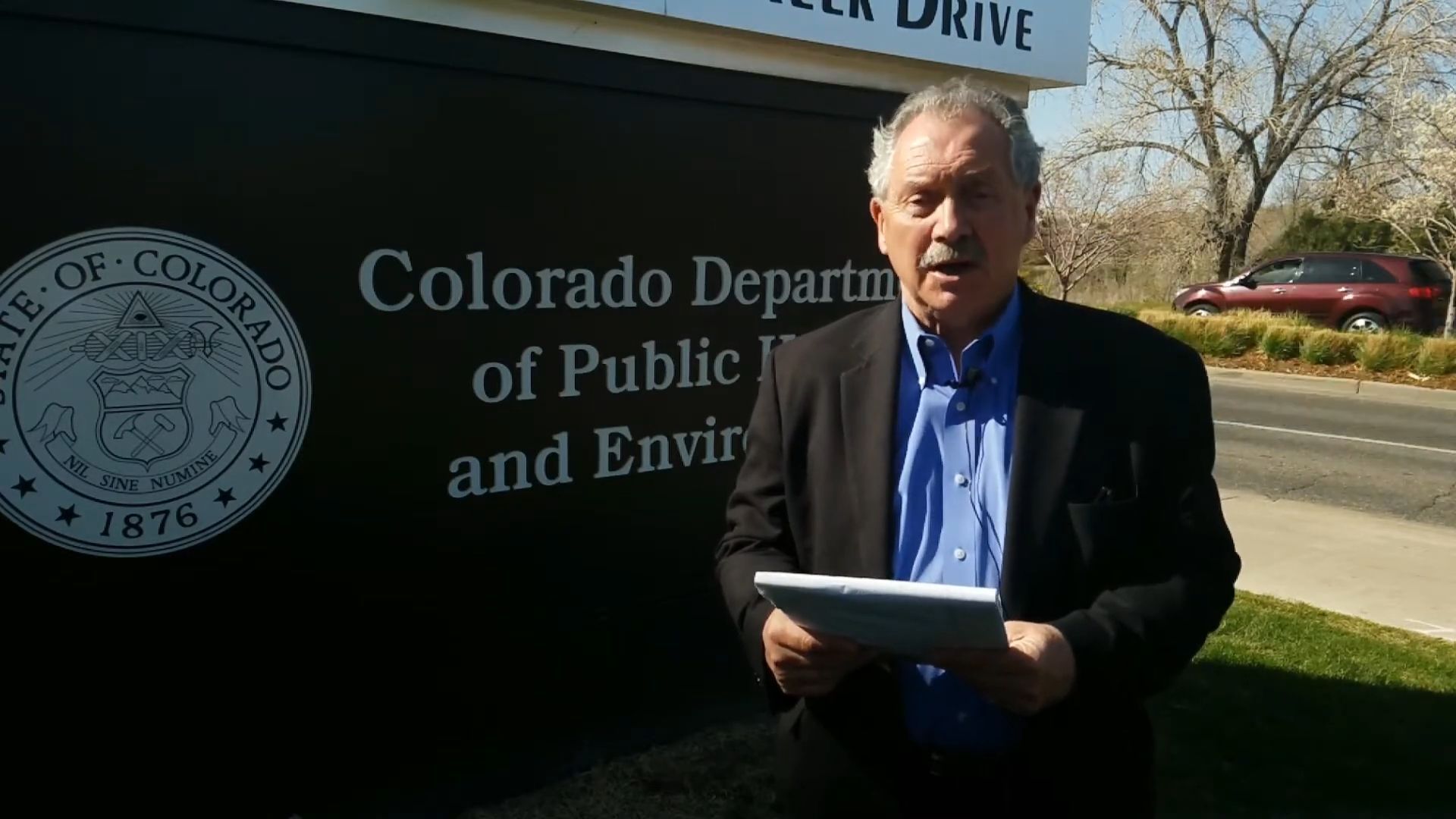 Man in a suit speaks outside the Colorado Department of Public Health and Environment building. He holds papers, and the sign is in view.