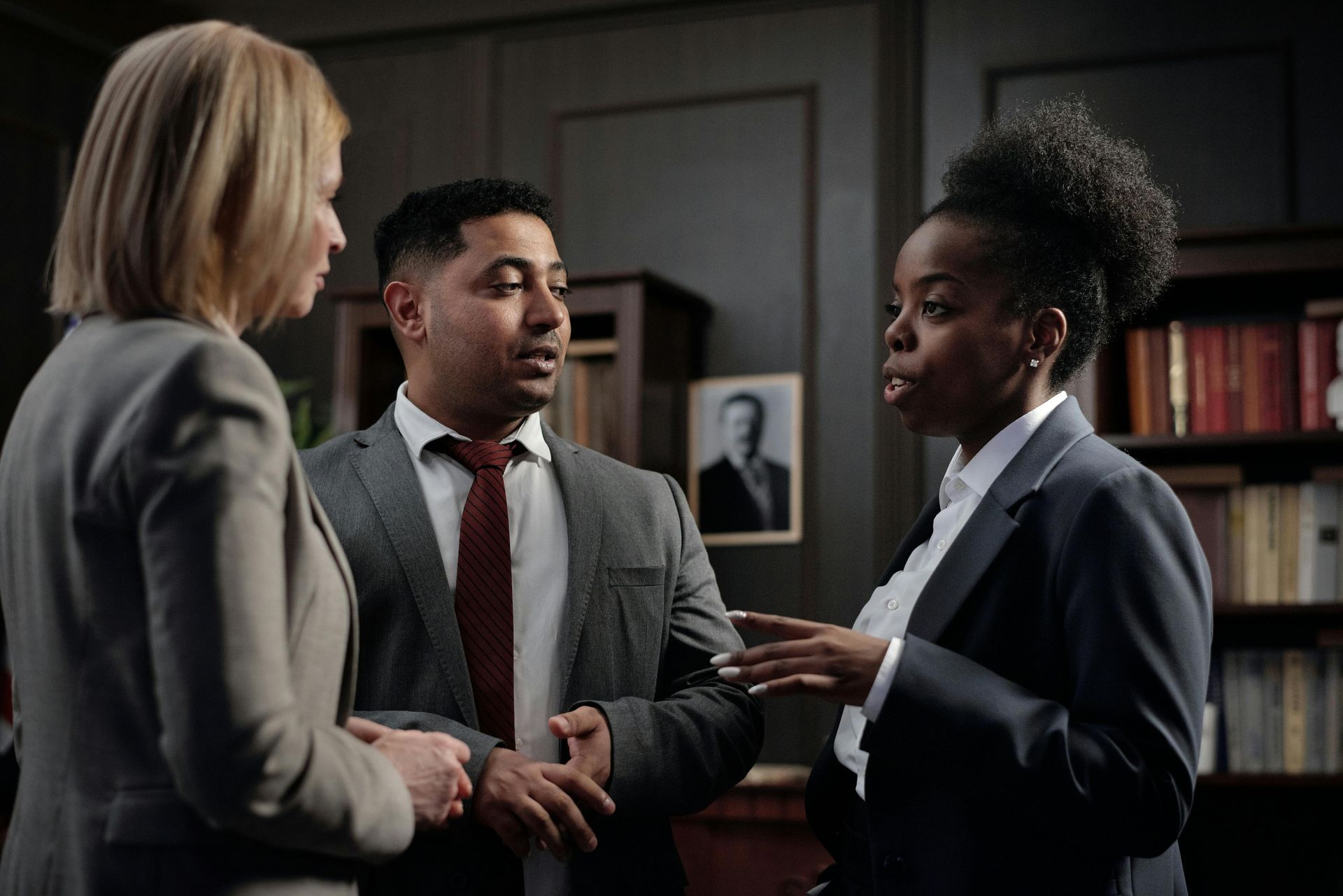 Three people in suits discussing in a library; one woman, a man, and another woman.