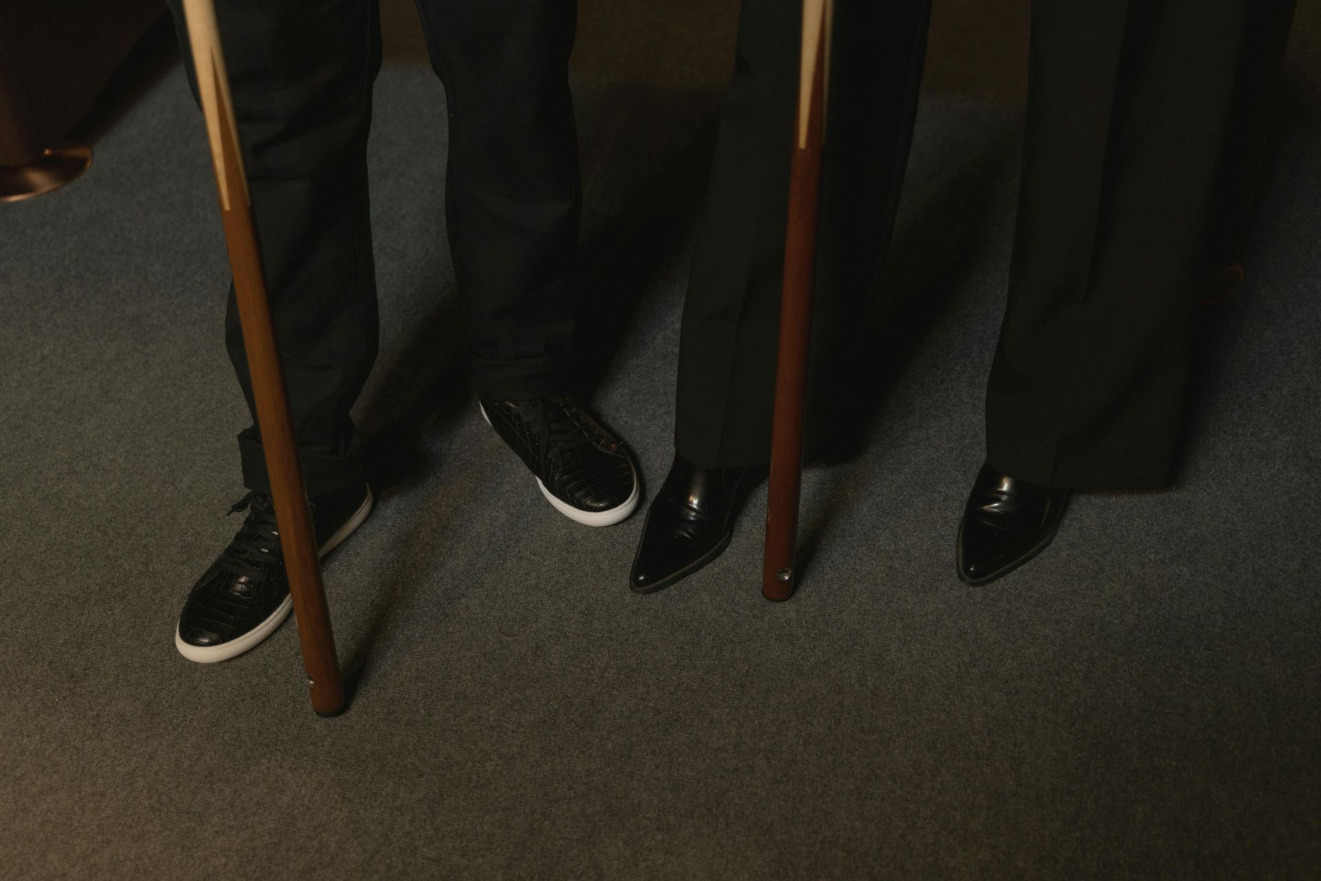 Four people's legs and pool cues standing on gray carpet. They are wearing black pants and shoes.