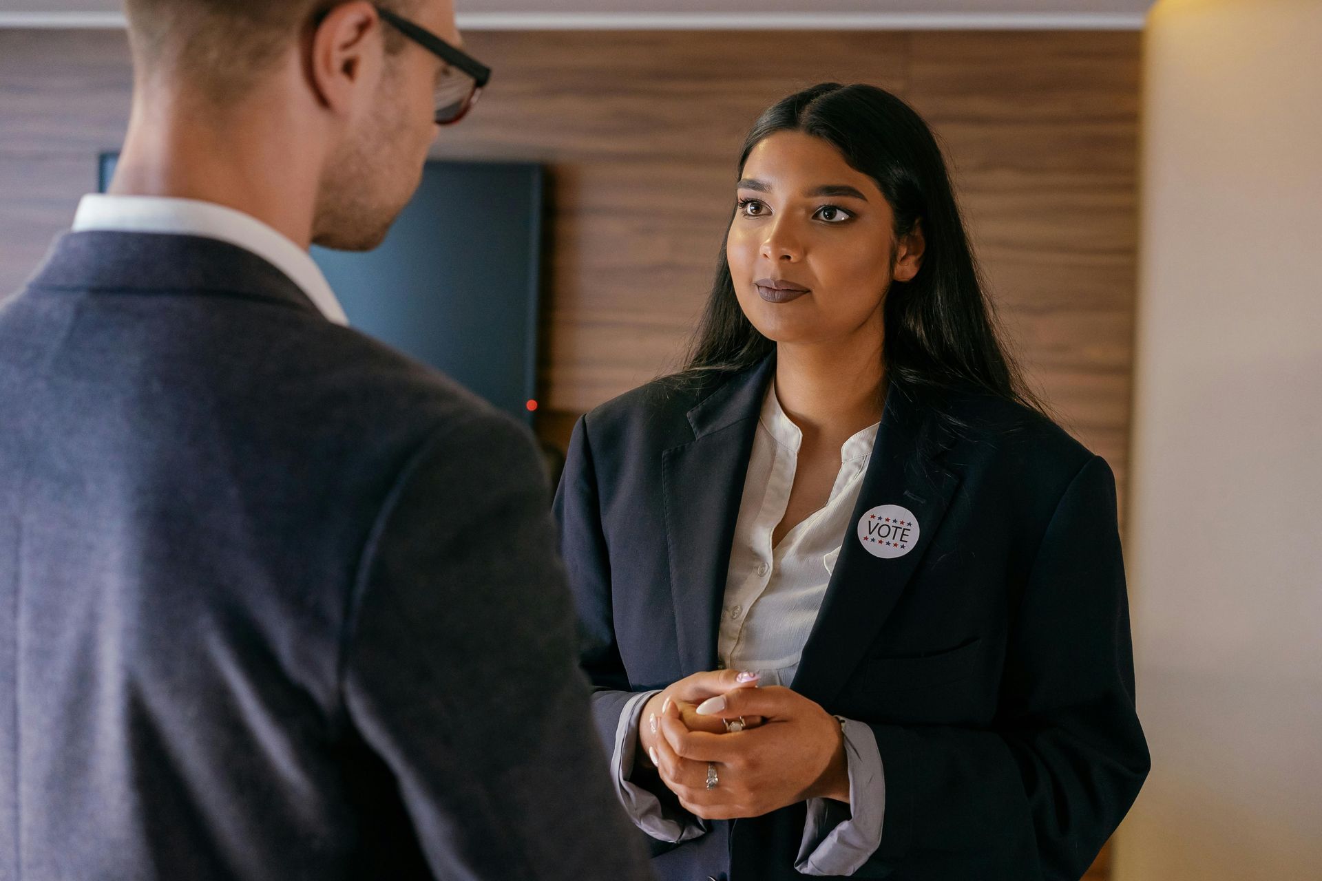 Woman in a blazer talks to a person wearing a suit. Dark wood paneling in background.