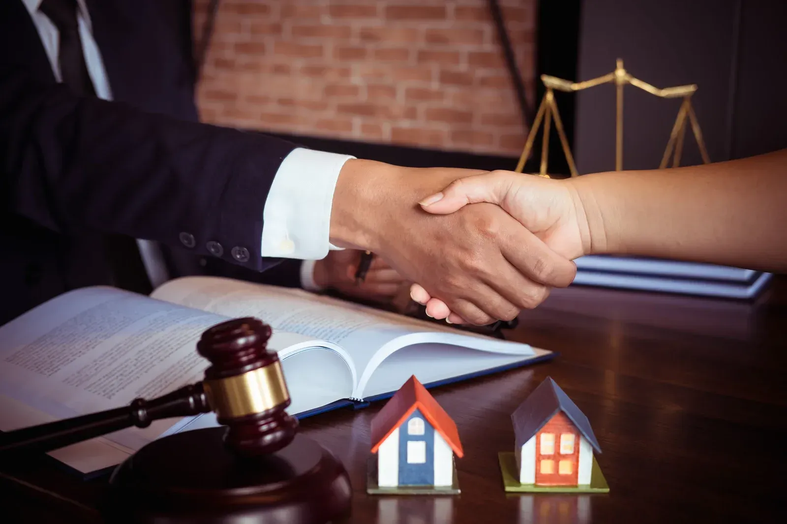 Two people shaking hands over a table with a gavel, open book, and house models.