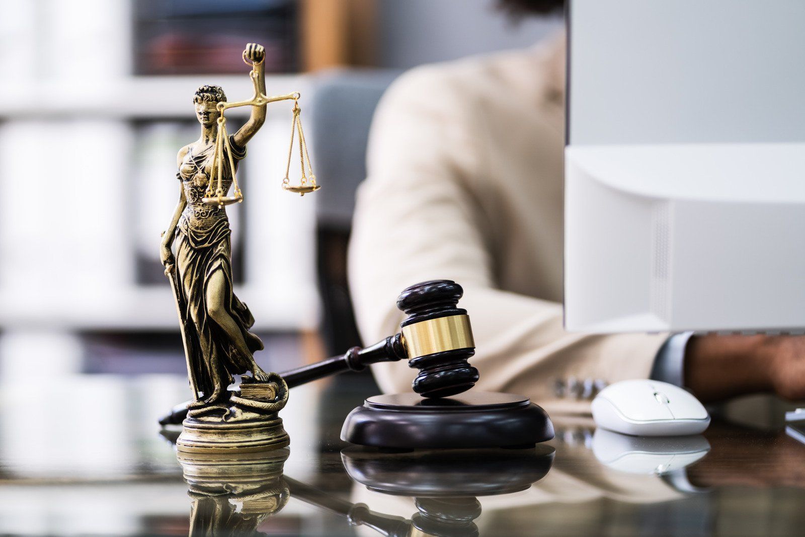 Statue of Lady Justice with scales and gavel on a desk with person using a computer in background.