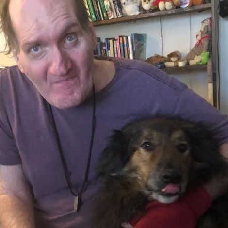 A man in a purple shirt is holding a brown dog in his arms.