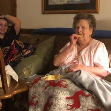 Two women are sitting on a couch eating food and drinking wine.