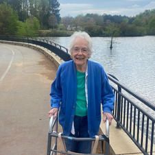 An elderly woman is walking with a walker on a path next to a lake.