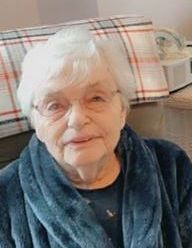 An elderly woman wearing glasses and a blue robe is sitting on a couch.