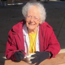 An elderly woman wearing a red jacket and black gloves is sitting at a table.