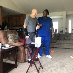 A nurse is helping an elderly man walk in a living room.