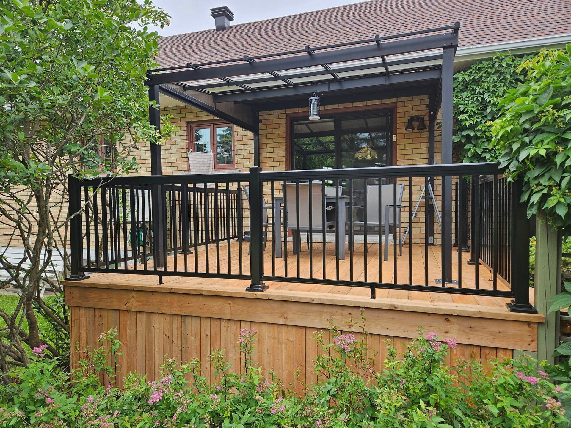 Une terrasse en bois avec une balustrade noire et un auvent au-dessus se trouve devant une maison.