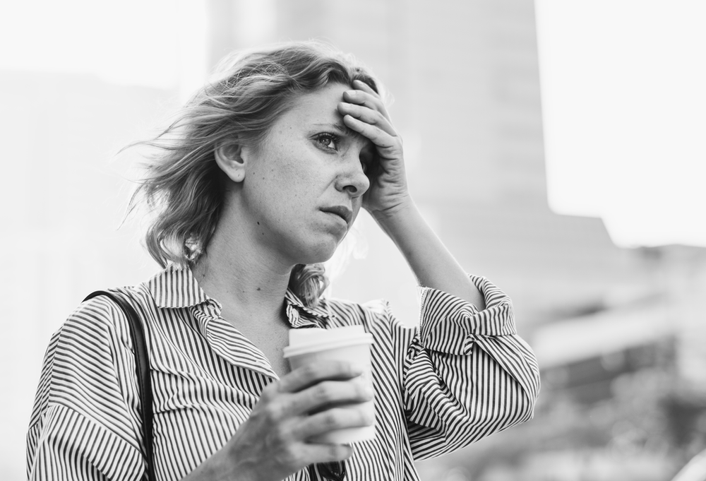 A woman is holding her hand to her forehead while holding a cup of coffee.