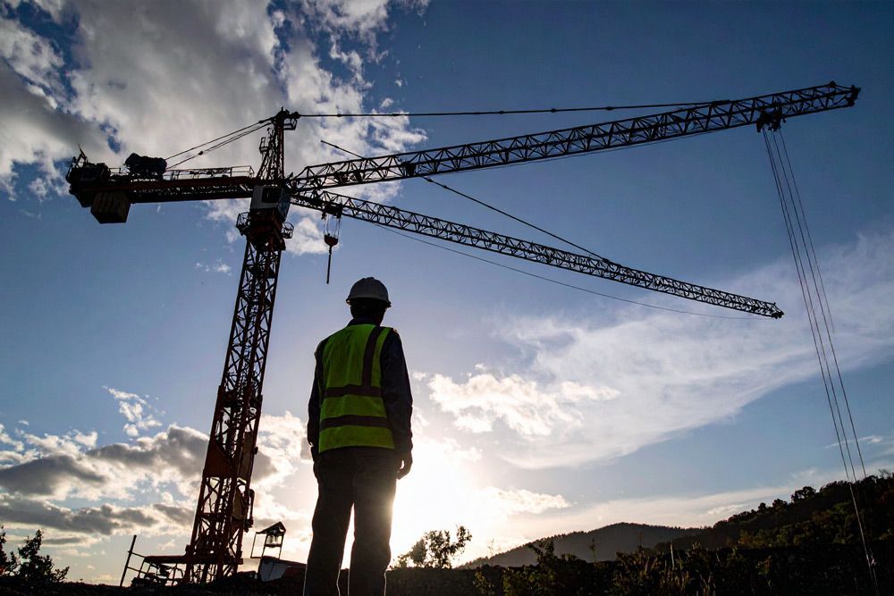 A construction worker is standing in front of a crane at sunset.