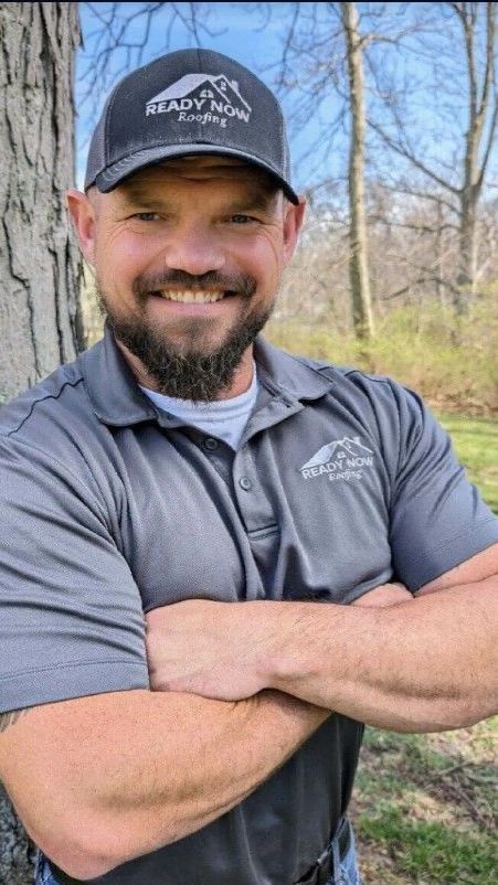 A man with a beard and a hat is standing with his arms crossed in front of a tree.