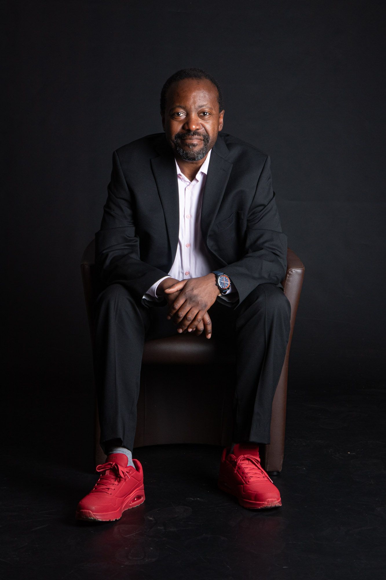 Man in a suit and red sneakers sits in a chair against a black backdrop, hands clasped.
