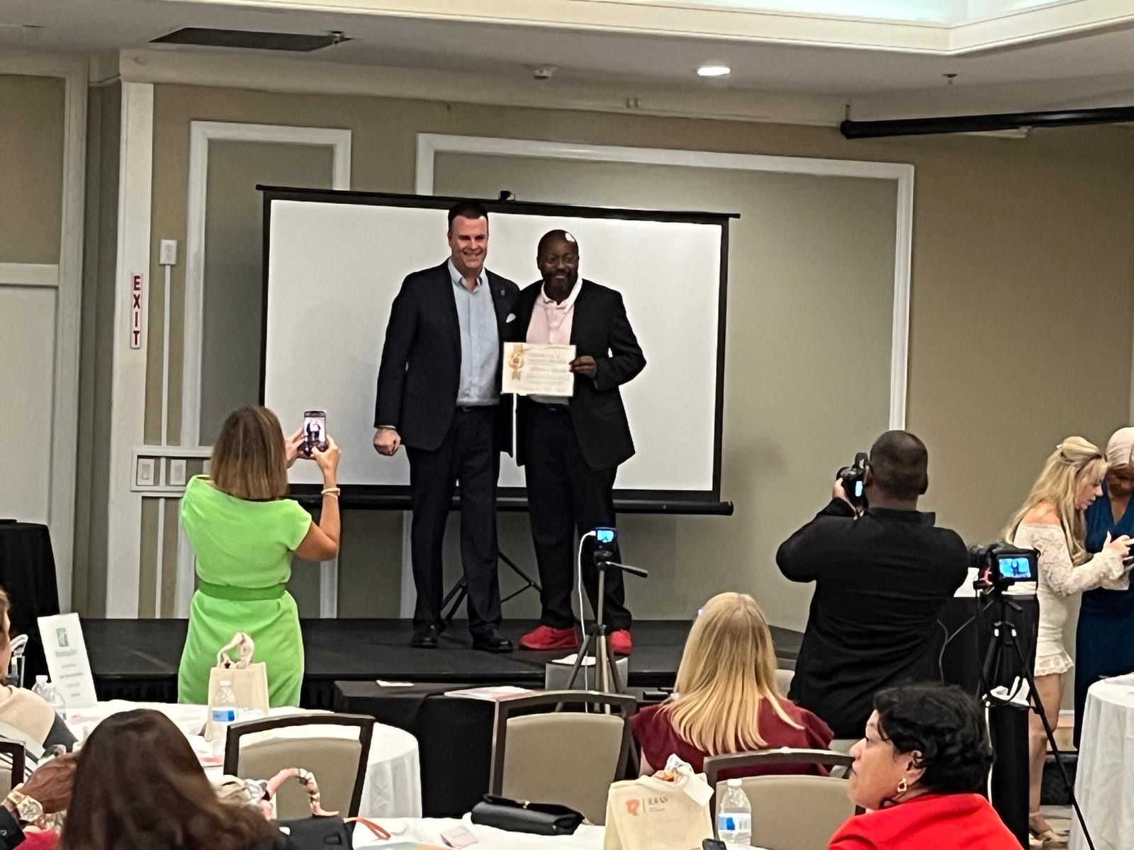 Two men stand on stage, one holding a certificate. People take photos, tables are set for a gathering.