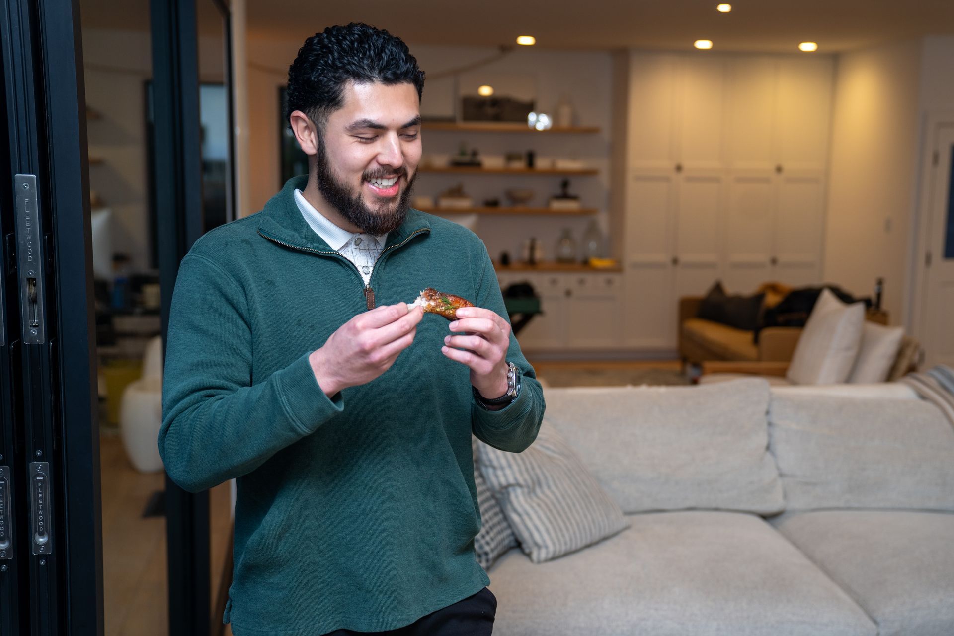 A man in a green sweater is eating a piece of food in a living room.