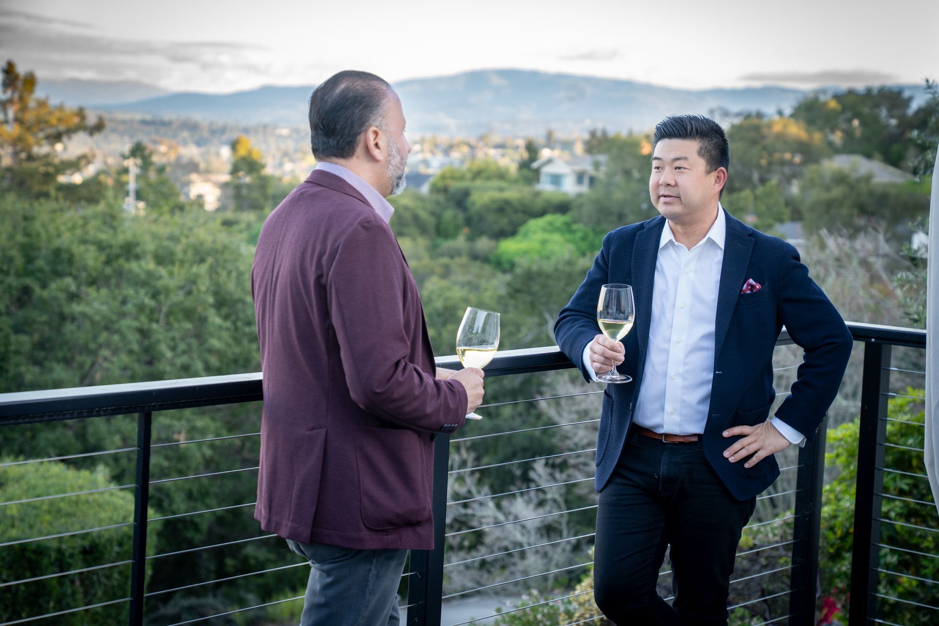 Two men are standing on a balcony holding wine glasses and talking.