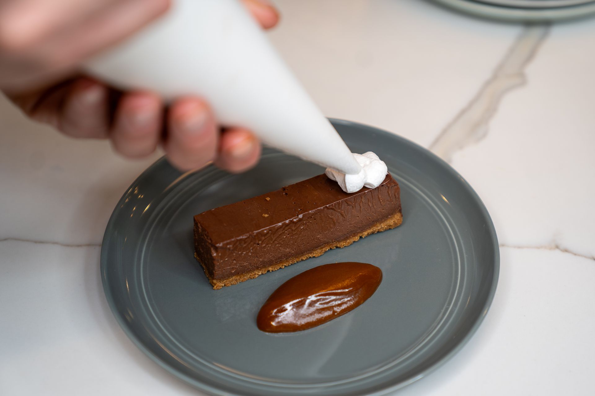 A person is icing a piece of chocolate cake on a plate.