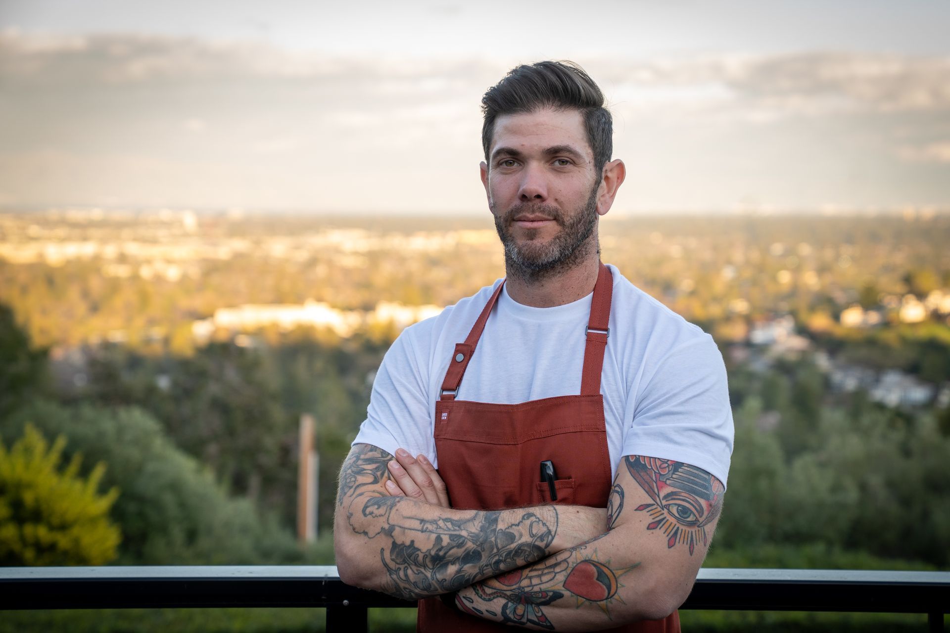 A man with tattoos is standing with his arms crossed on a balcony.
