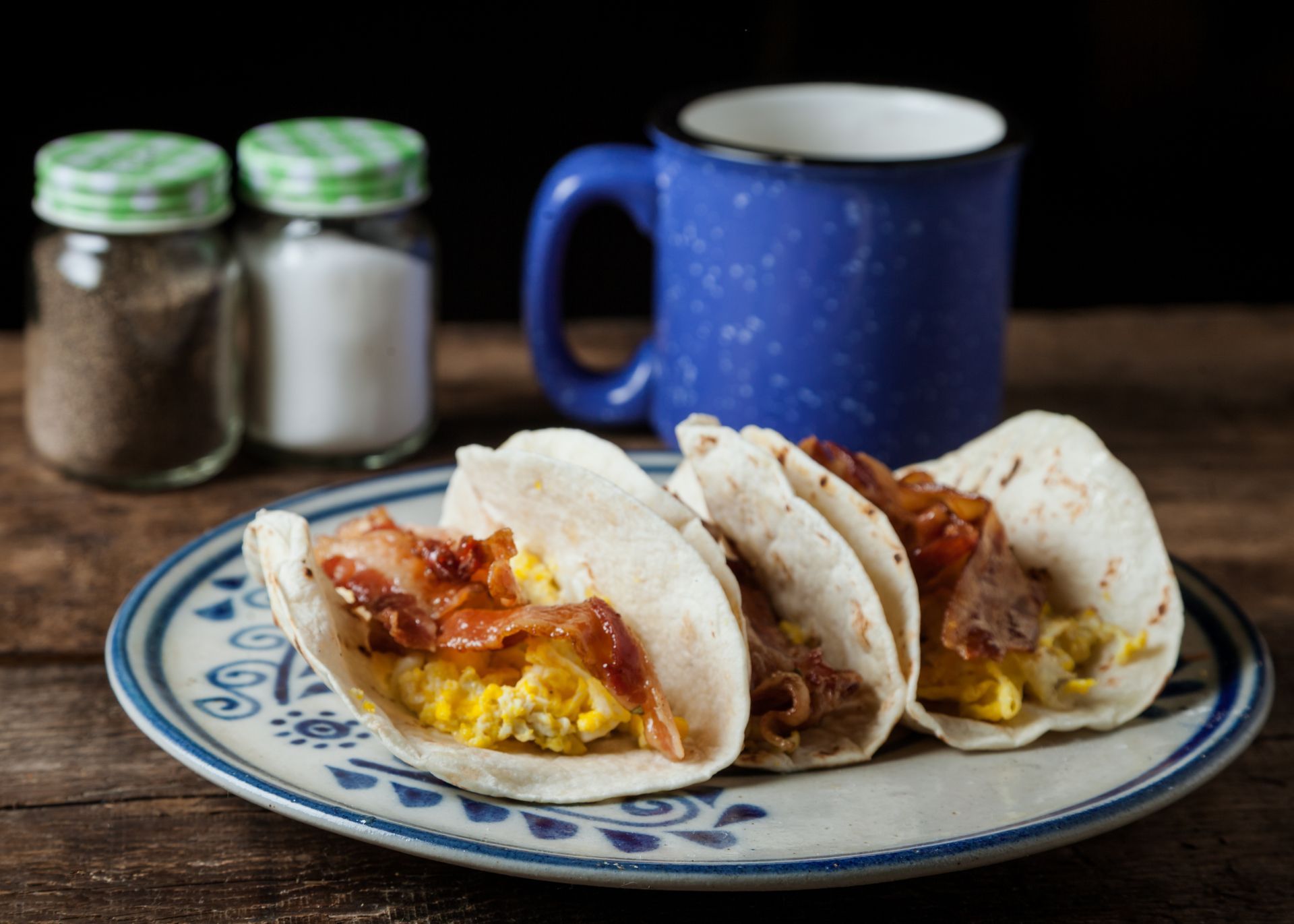 Three tacos with bacon and eggs on a plate next to a cup of coffee.