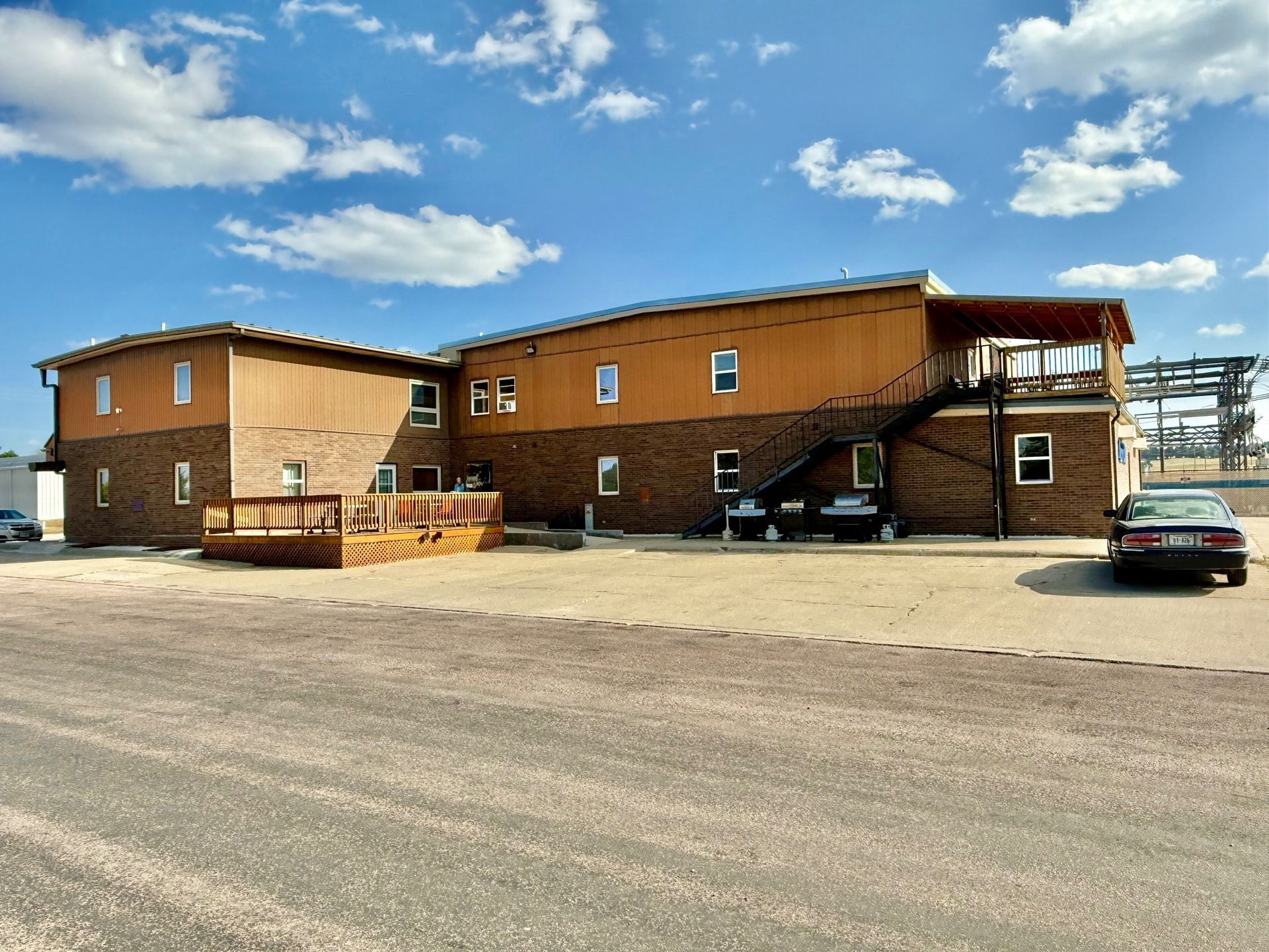 Two-story brick and wood building with multiple windows under a blue sky, gravel parking lot.