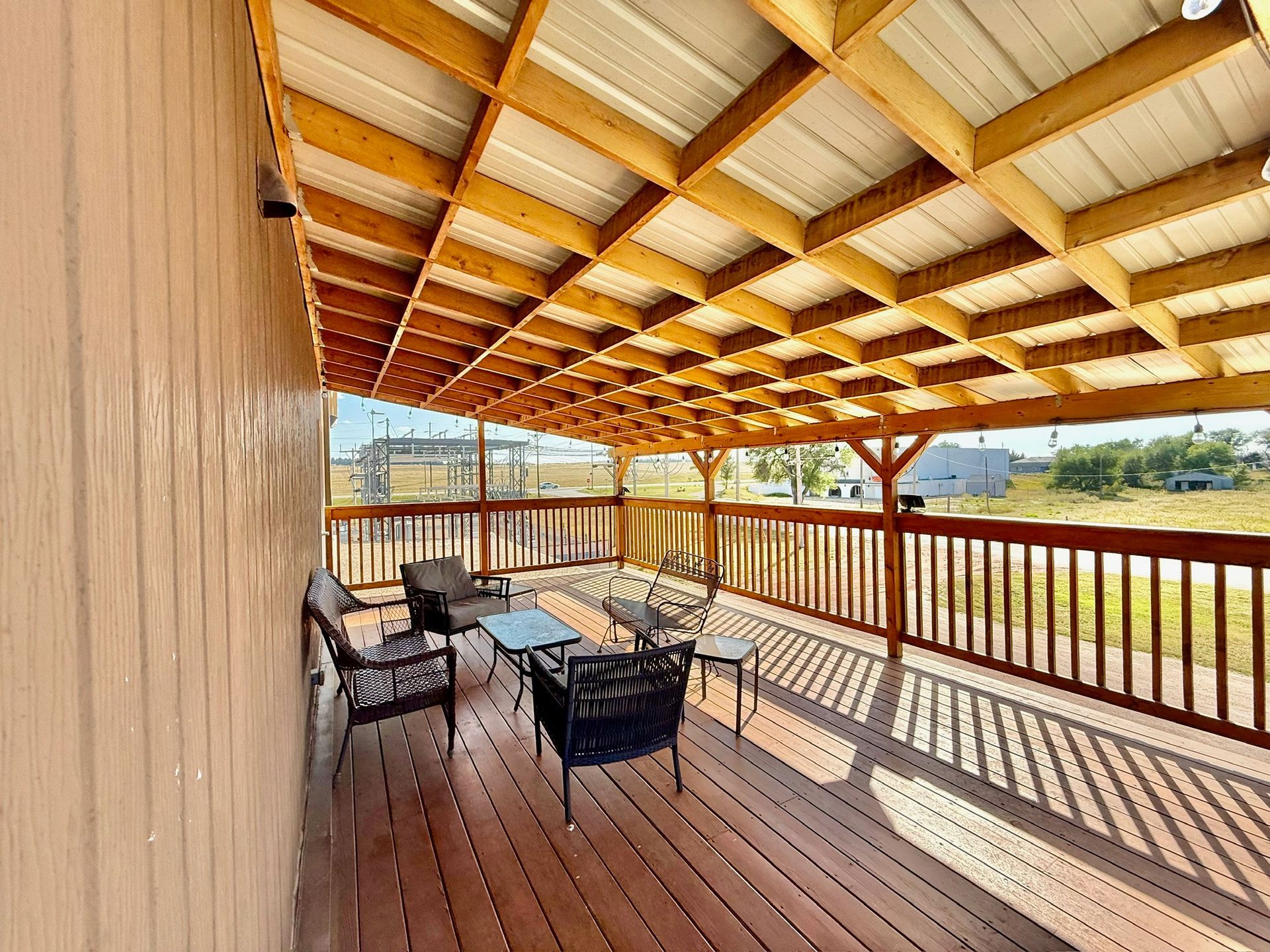 Wooden deck with seating under a wood and metal roof, viewed from the side of a building.