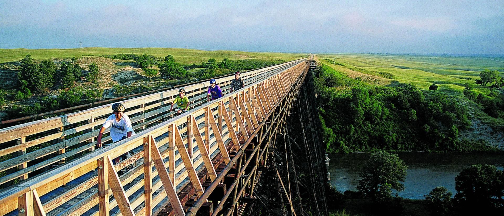 People walking on a long wooden bridge over a river, surrounded by green fields and trees under a blue sky.