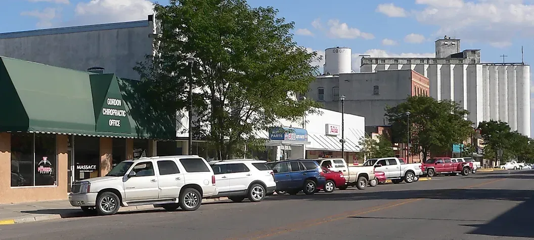 Street scene with parked cars, buildings, and grain elevators in the background on a sunny day.