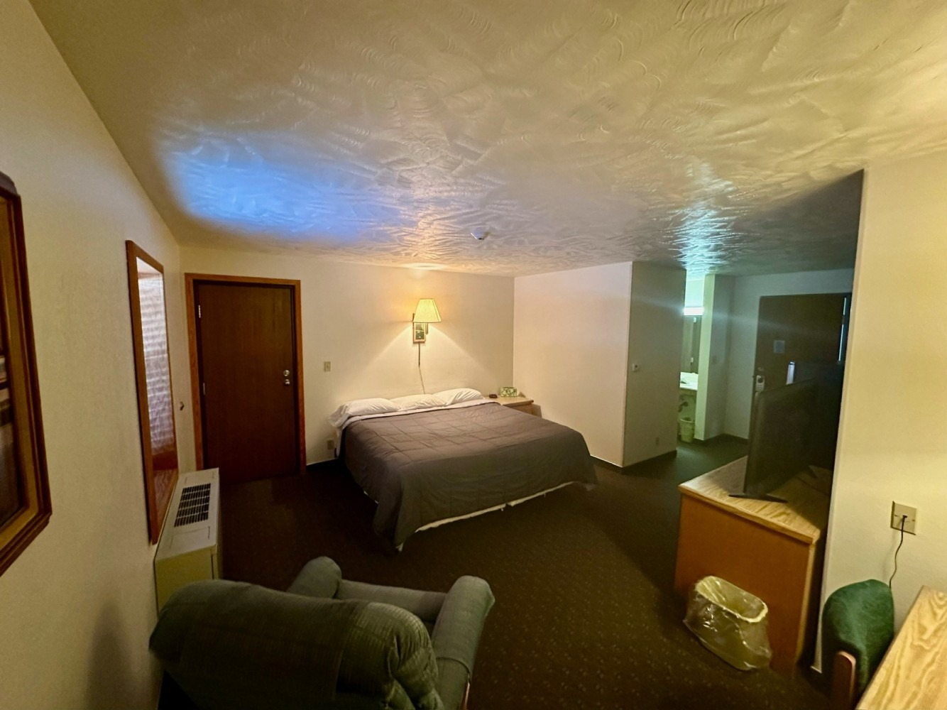 Motel room with bed, chair, desk, and bathroom doorway. Dark brown carpet, light walls.