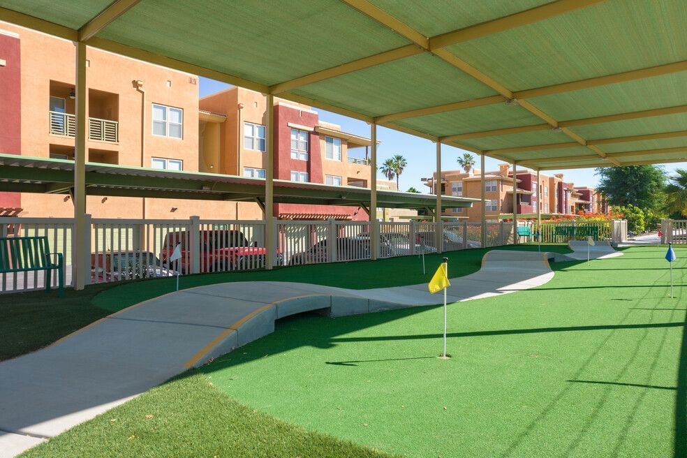 Miniature golf course covered by a green shade structure. Course has green turf, concrete paths, and yellow flags. Apartment buildings are in the background.