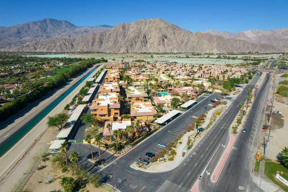 Aerial view of a residential community in a desert landscape, featuring rows of buildings, a highway, a canal, and mountains in the background.