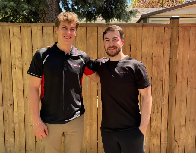 Two smiling men standing outdoors in front of a wooden fence, one in a black polo and one in a black T-shirt.