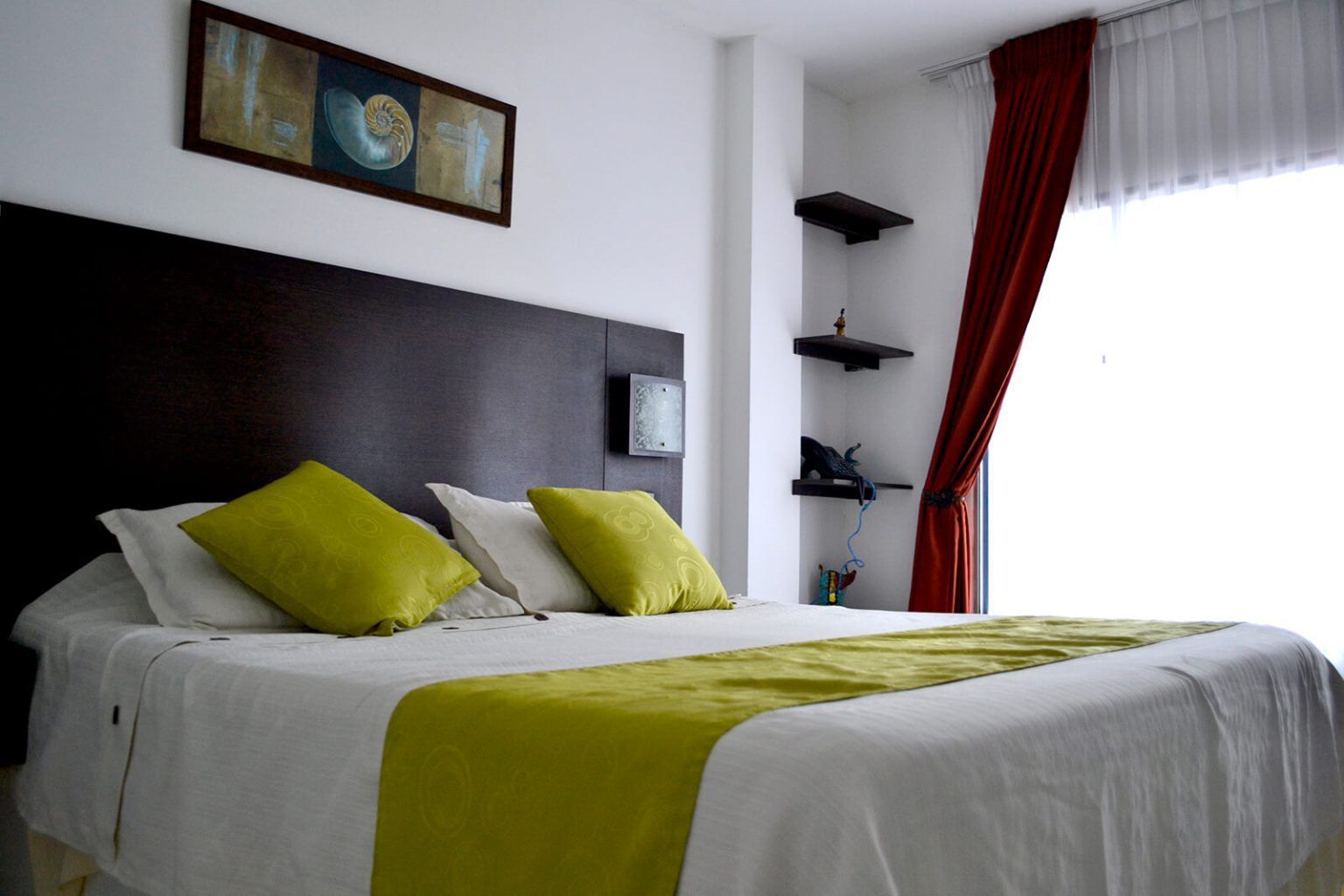 Dormitorio moderno con cabecero oscuro, ropa de cama verde, cuadros en la pared y cortinas rojas junto a una ventana luminosa.