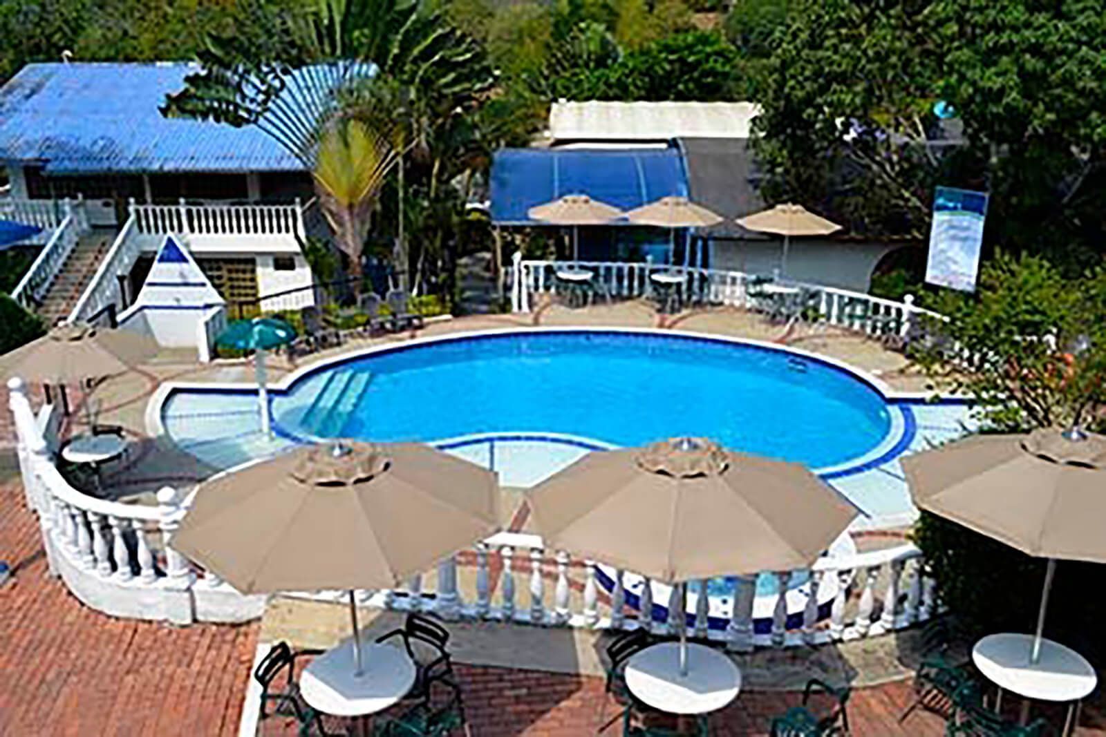 Piscina exterior de un complejo turístico con agua azul, barandilla blanca, sombrillas color canela y tumbonas bajo un cielo soleado.