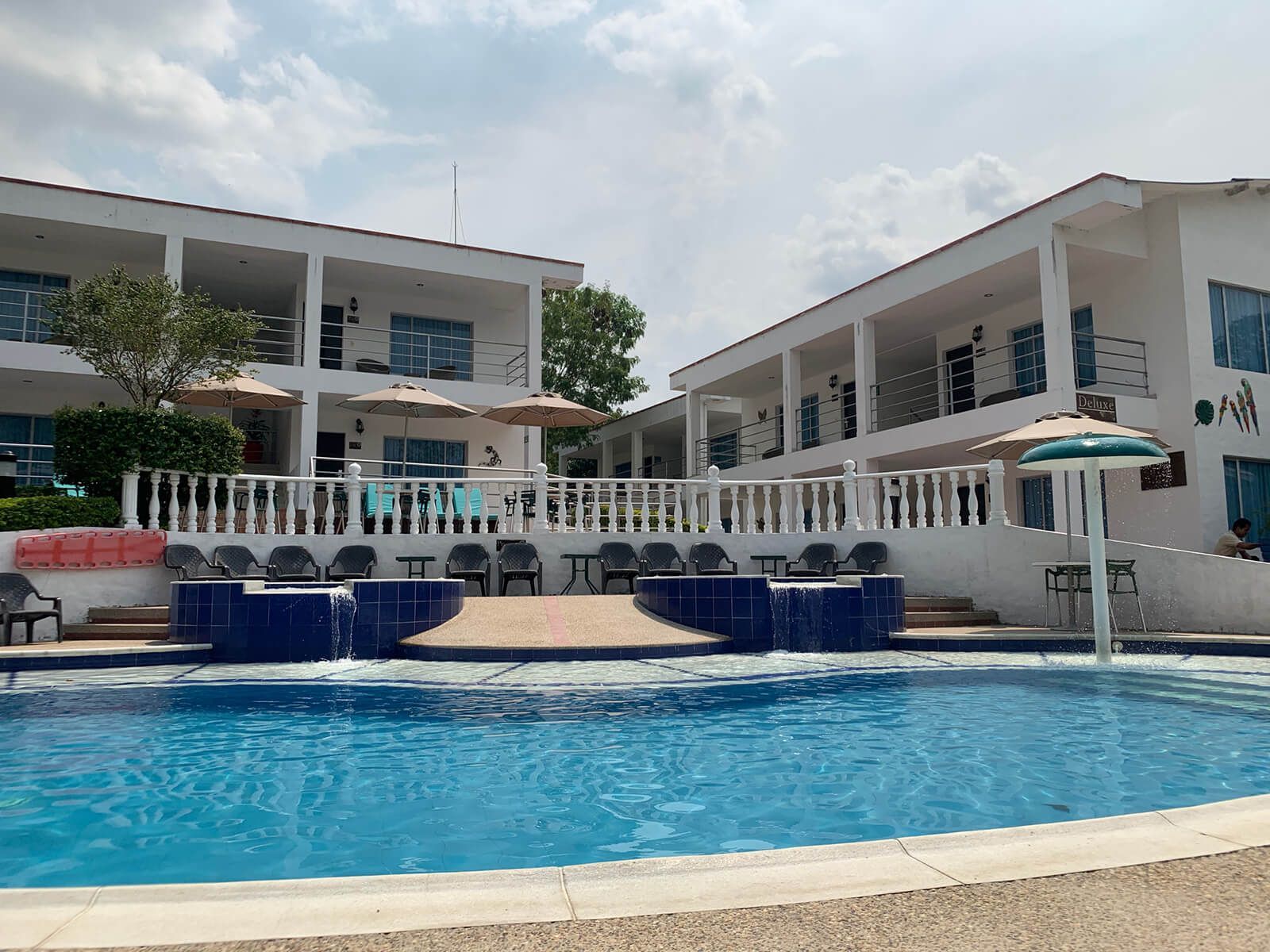 Piscina del complejo turístico con agua azul, edificios blancos y tumbonas bajo un cielo parcialmente nublado.