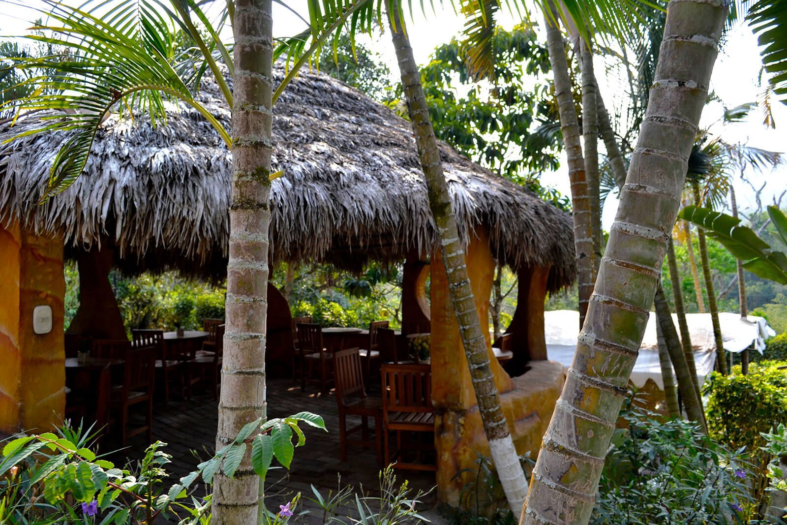 Cabaña tropical en la playa con techo de paja y palmeras, con vistas a las tumbonas junto a la orilla.