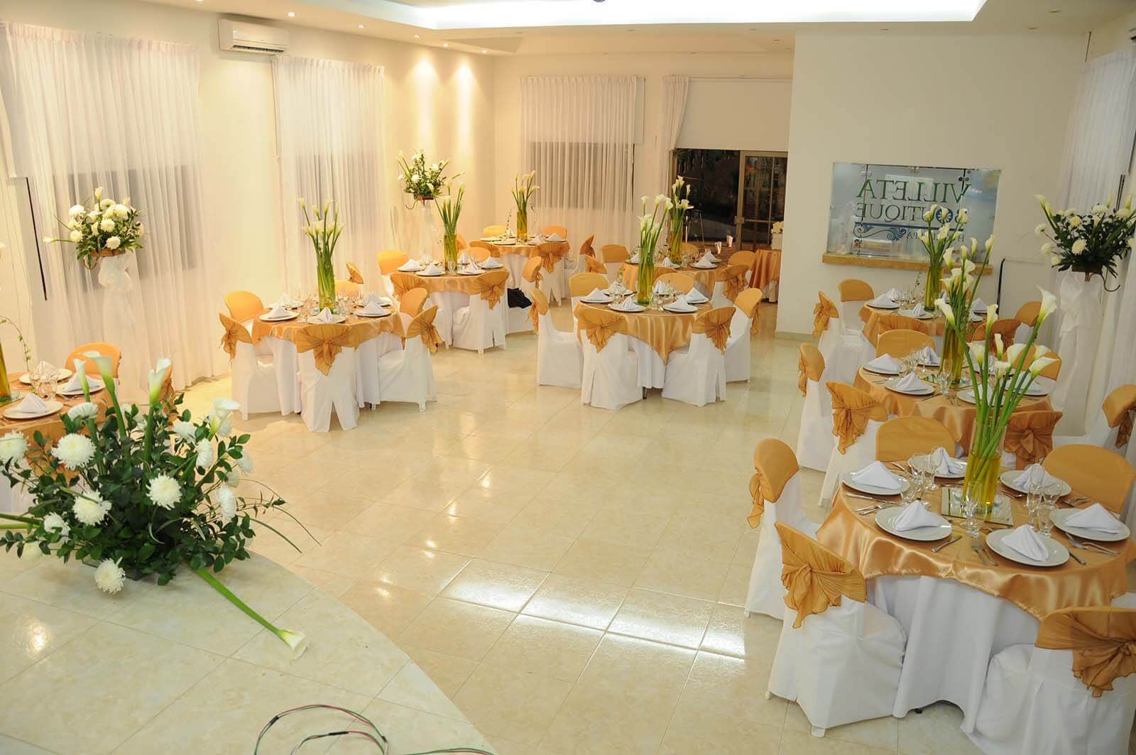 Salón de banquetes decorado con mesas redondas, sillas blancas y detalles dorados, preparado para un evento.