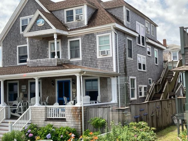 Gray shingle-clad multi-story house with porch, side stairs, and flowers in front.