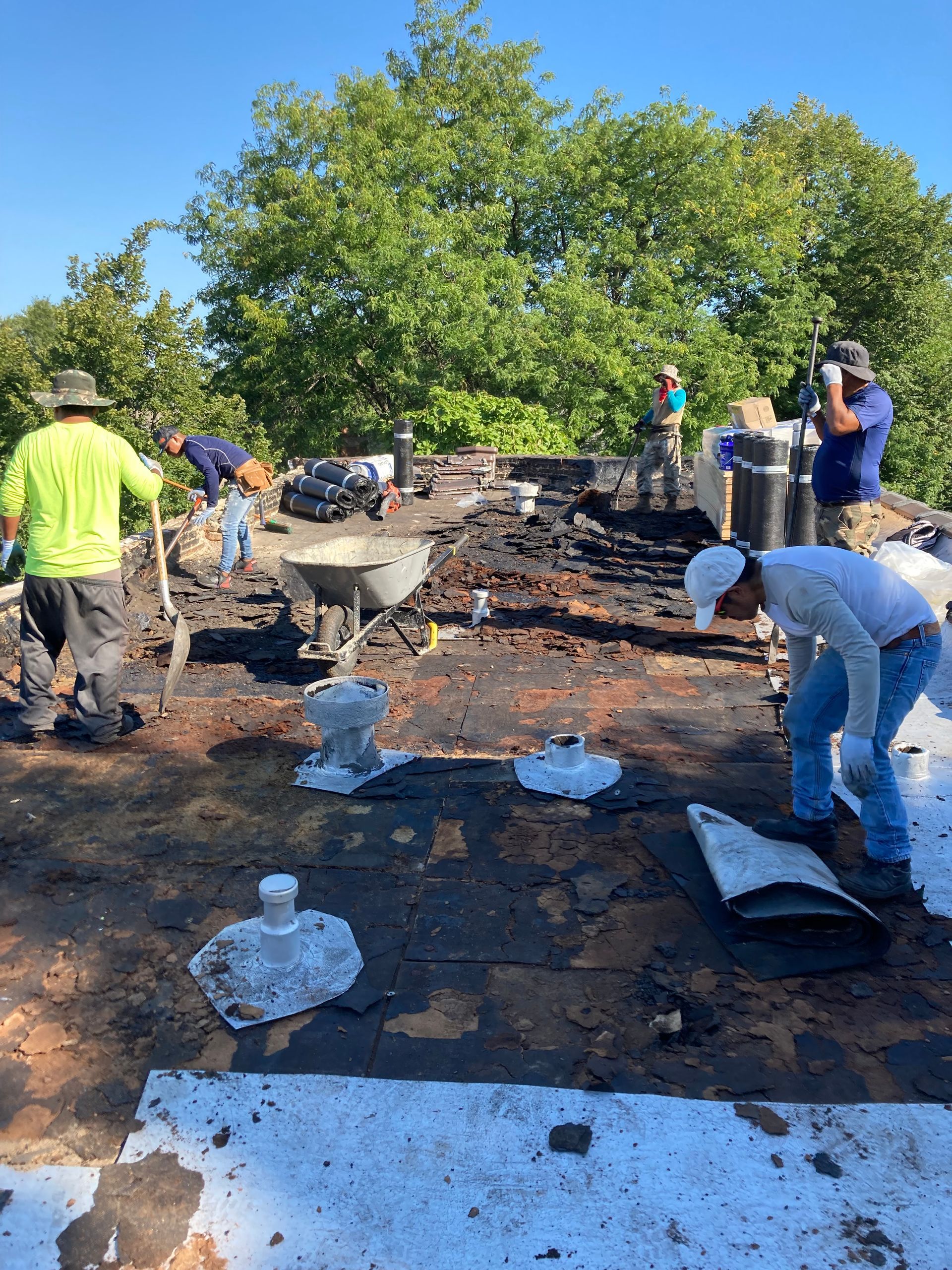 Construction workers repairing a roof. People are using tools, with a wheelbarrow present. Blue sky, trees in the background.